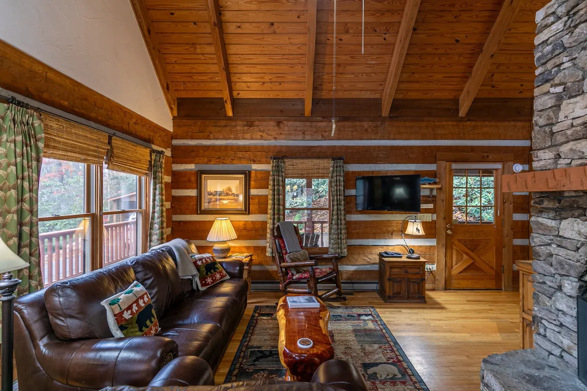 An inviting living space with a leather sofa, stone fireplace, wooden beams, television, and artwork on the wall. Large windows offer nature views, enhancing the connection to the outdoors while inside. The setting is ideal for streaming movies, relaxing with a book, or sharing stories by the fire.
