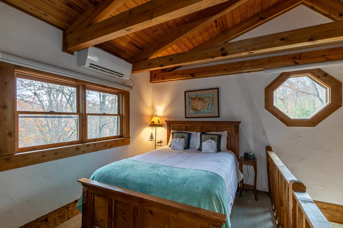 A cozy loft area with a queen bed positioned beneath exposed wooden beams, adorned with warm bedding and a lamp on the nightstand. Windows, including a unique octagonal one, bring in natural light, creating a sense of openness and comfort perfect for families or group sleepovers.