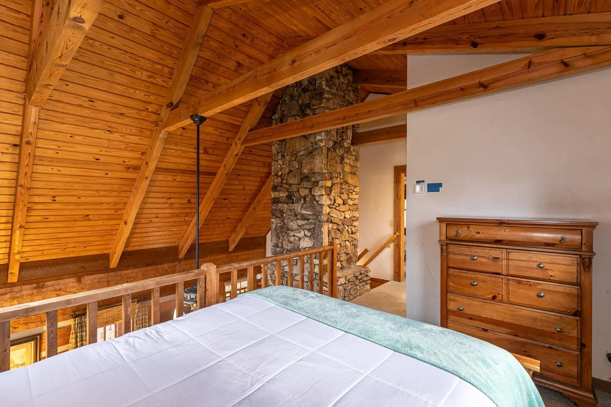 This image captures the cozy interior of the log cabin, with rustic wooden beams, a striking stone fireplace, and an antique dresser. The inviting furniture suggests a warm place to relax after a day of outdoor adventure, and the balcony hints at the surrounding natural beauty.