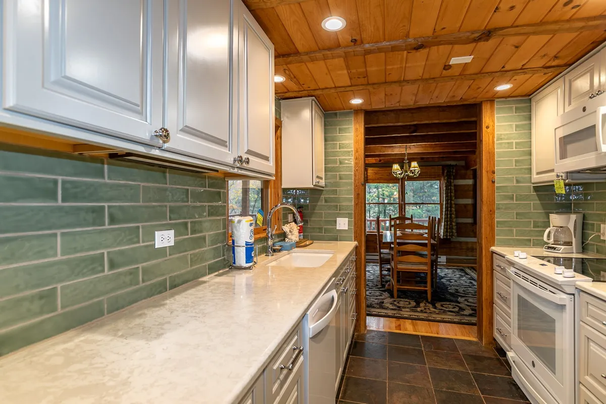 A warm, inviting kitchen with a wooden counter, green tile backsplash, exposed beams, and a view into the dining area beyond. The setup signals a communal space ideal for preparing group meals and gathering for breakfast before a day of adventure.