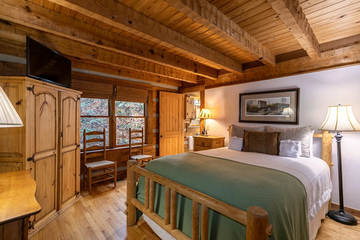 A tranquil bedroom scene featuring a queen bed, wooden furniture, and large windows that invite in soft daylight. The room feels peaceful and perfect for relaxation or reading, with a lamp on the nightstand and simple, calming decor.