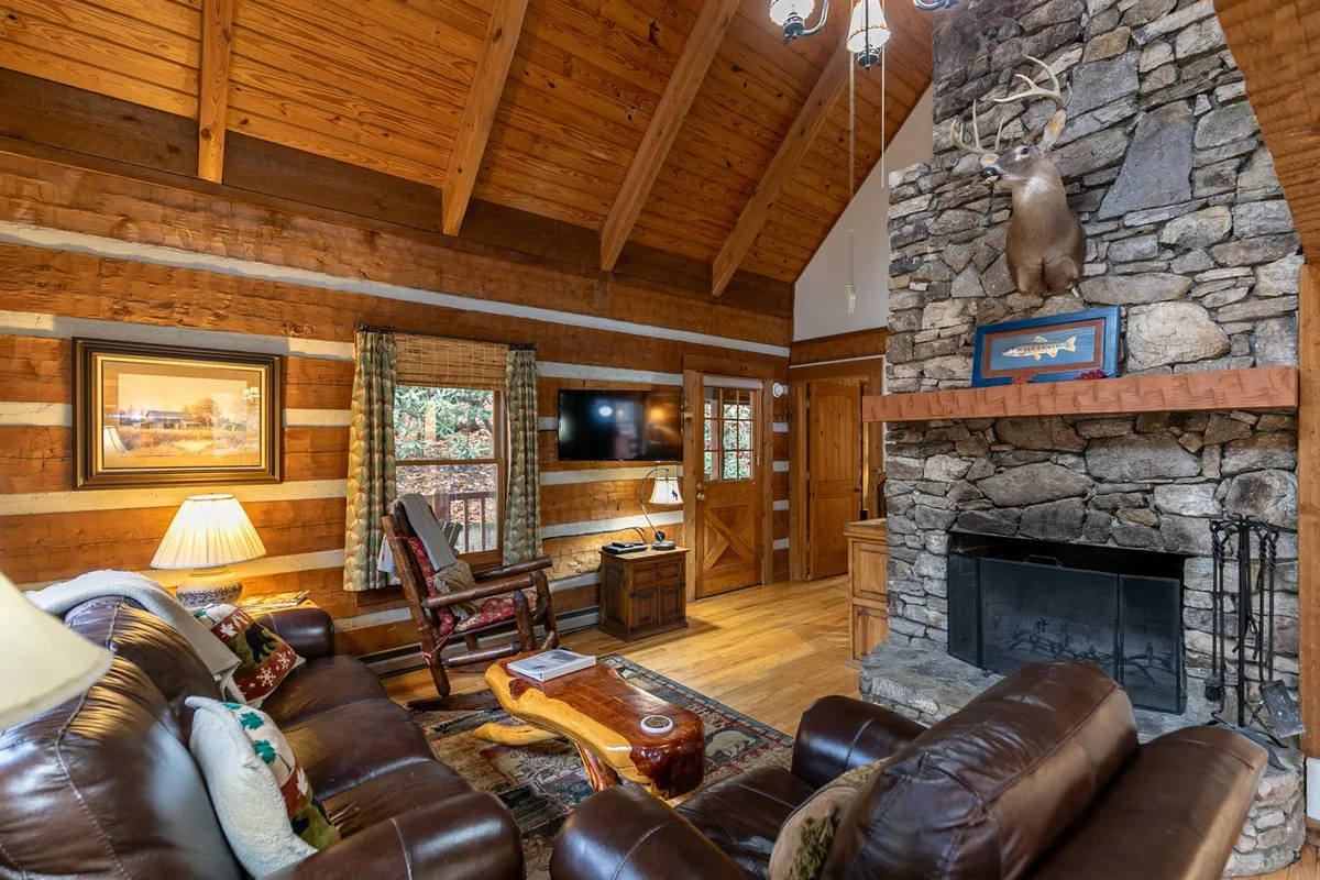 A spacious living area is shown with a prominent stone fireplace, leather sofas, exposed wooden beams, and tasteful rustic decor. The room feels inviting and warm, ideal for relaxing by the fire, streaming movies, or sharing laughter with loved ones. The decorative touches and soft lighting enhance the sense of comfort and togetherness.