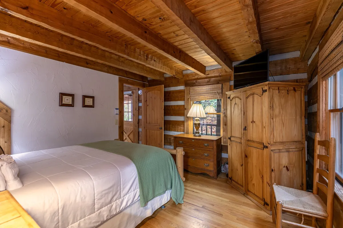 A warmly lit bedroom with wooden beams overhead, a queen bed dressed in white linens, a sturdy wooden nightstand, and a lamp. Natural light pours in through the window beside the bed, illuminating the cozy wooden furniture and creating a peaceful atmosphere.