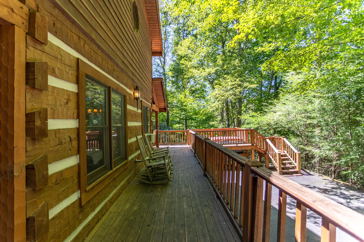 A wide view of the wrap-around wooden deck nestled among mature trees, featuring rustic rocking chairs strategically placed for relaxing while enjoying the mountain air. The deck's elevated position provides a peaceful vantage point over the natural two-acre wooded setting.