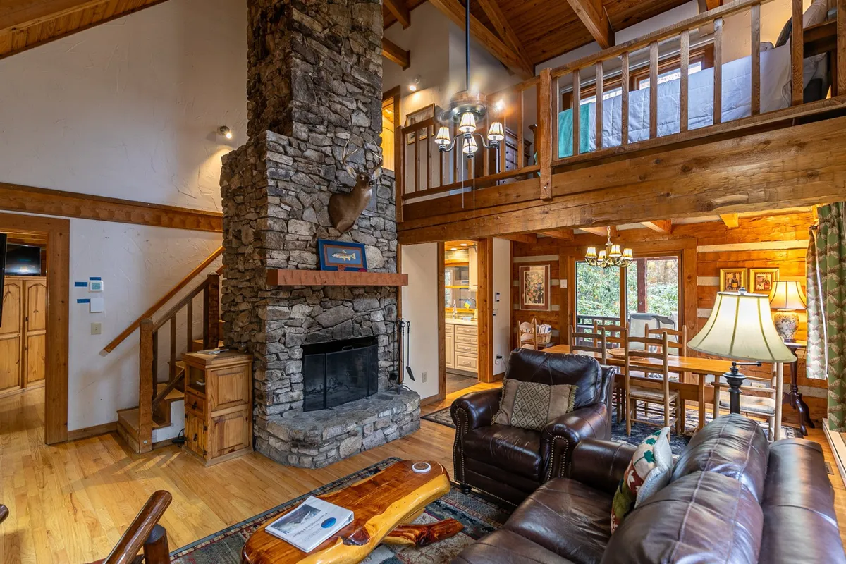 A wide view of the central living area, featuring a dramatic stacked stone wood-burning fireplace, soaring wooden beams, rich hardwood floors, and comfortable leather seating. The adjoining dining area is visible, creating a sense of connectedness between the living and dining spaces.