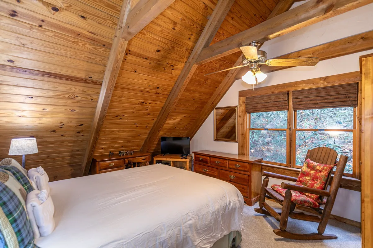 A rocking chair is positioned by a large window, framed by wooden ceiling beams, offering a peaceful spot to relax and connect with the outdoors. Through the window, natural light and hints of the wilderness beyond invite guests to enjoy the scenery.
