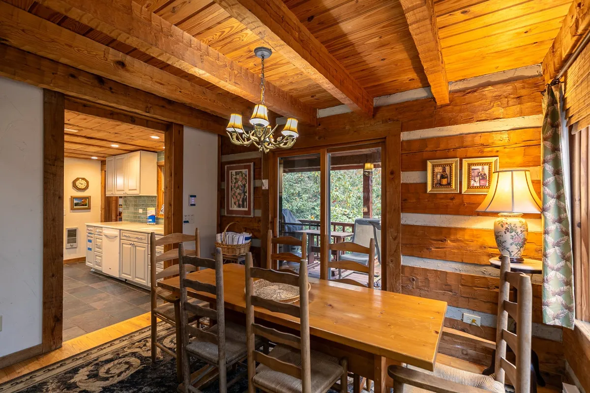 A close-up of the spacious dining area, highlighting the hand-made wooden table set for six beneath a striking deer antler chandelier. Exposed wooden beams and large windows bring in natural light, amplifying the warmth and rustic elegance.