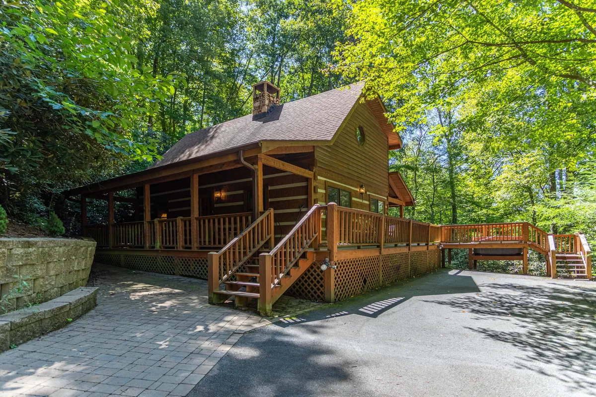 The image showcases a charming log cabin nestled among tall trees, with a spacious wooden deck overlooking two acres of private, wooded surroundings. The warm glow from the windows invites guests to imagine cozy nights inside, while the expansive deck hints at sunset gatherings and outdoor relaxation amidst nature.