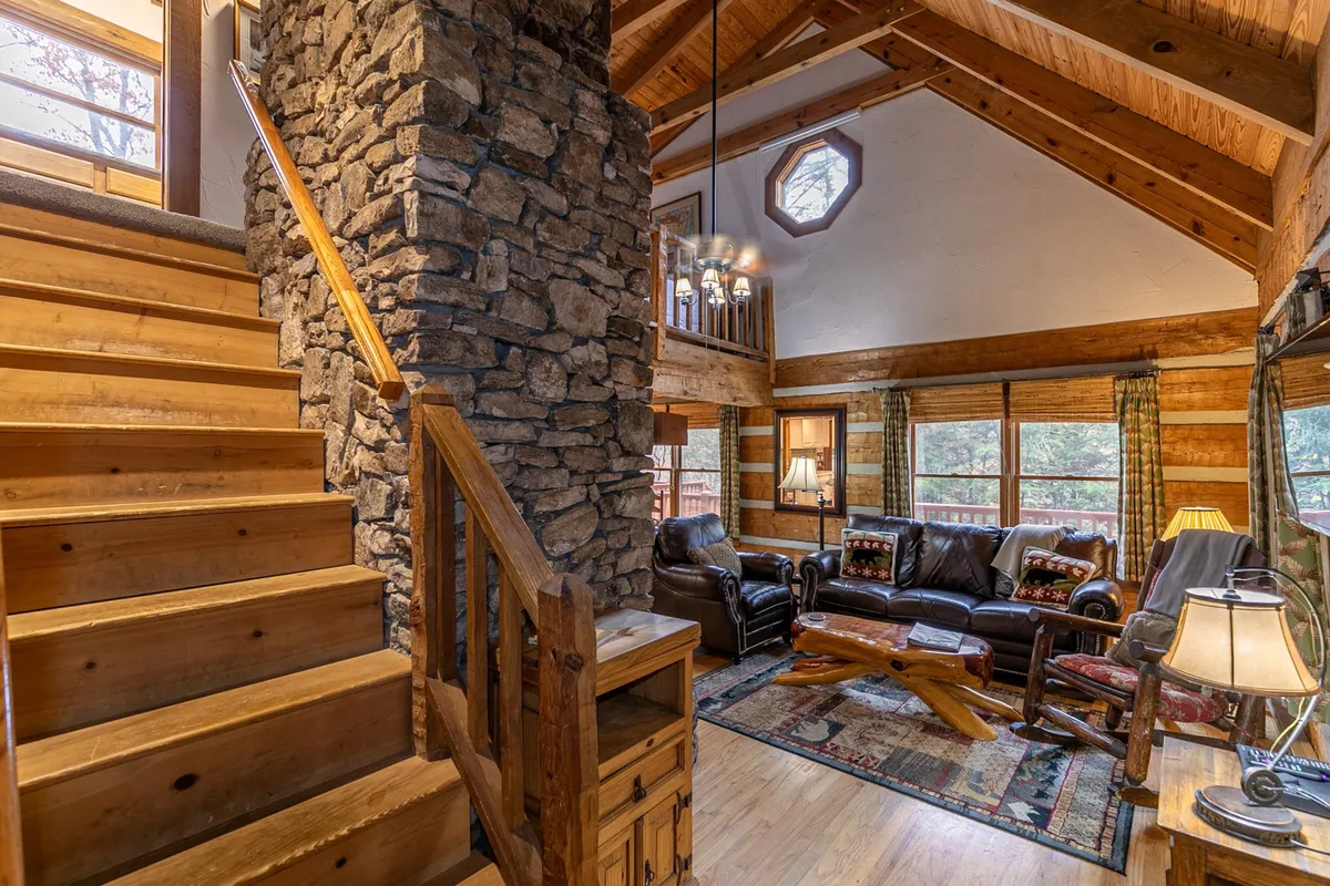 A cozy living room framed by a stone fireplace, wooden staircase, leather couches, and an octagonal window with natural light streaming in. The space feels inviting and is ideal for group conversations, game nights, or relaxing by the fire after adventuring.