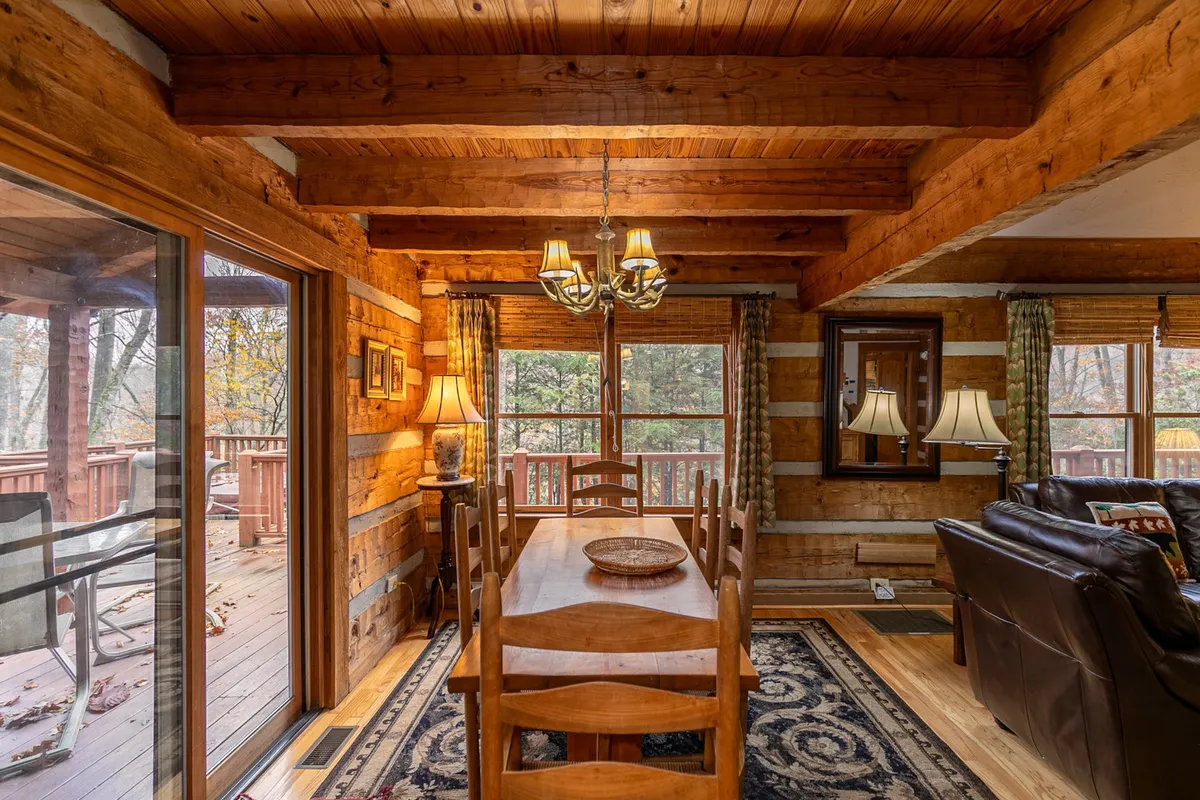 A stylish dining area with a large wooden table under a rustic chandelier, featuring prominent windows that connect the space to the outdoors. The scene is set for group dinners, intimate meals, or game nights in a naturally bright, social environment.