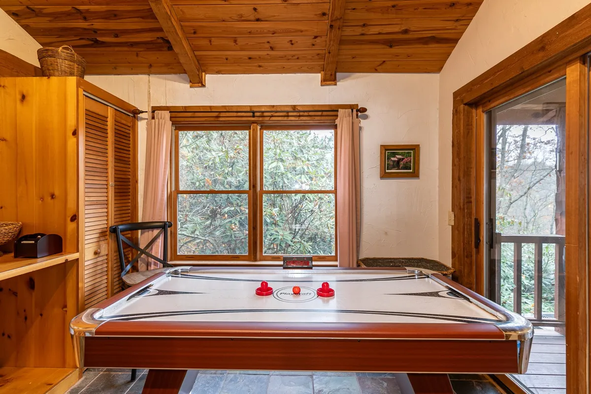 This image features an air hockey table positioned in front of large windows that invite natural light into the room. Wooden cabinetry and rustic touches frame the space, while the view outside offers a scenic backdrop. The room exudes a playful, relaxed energy, perfect for friendly competition and group entertainment.